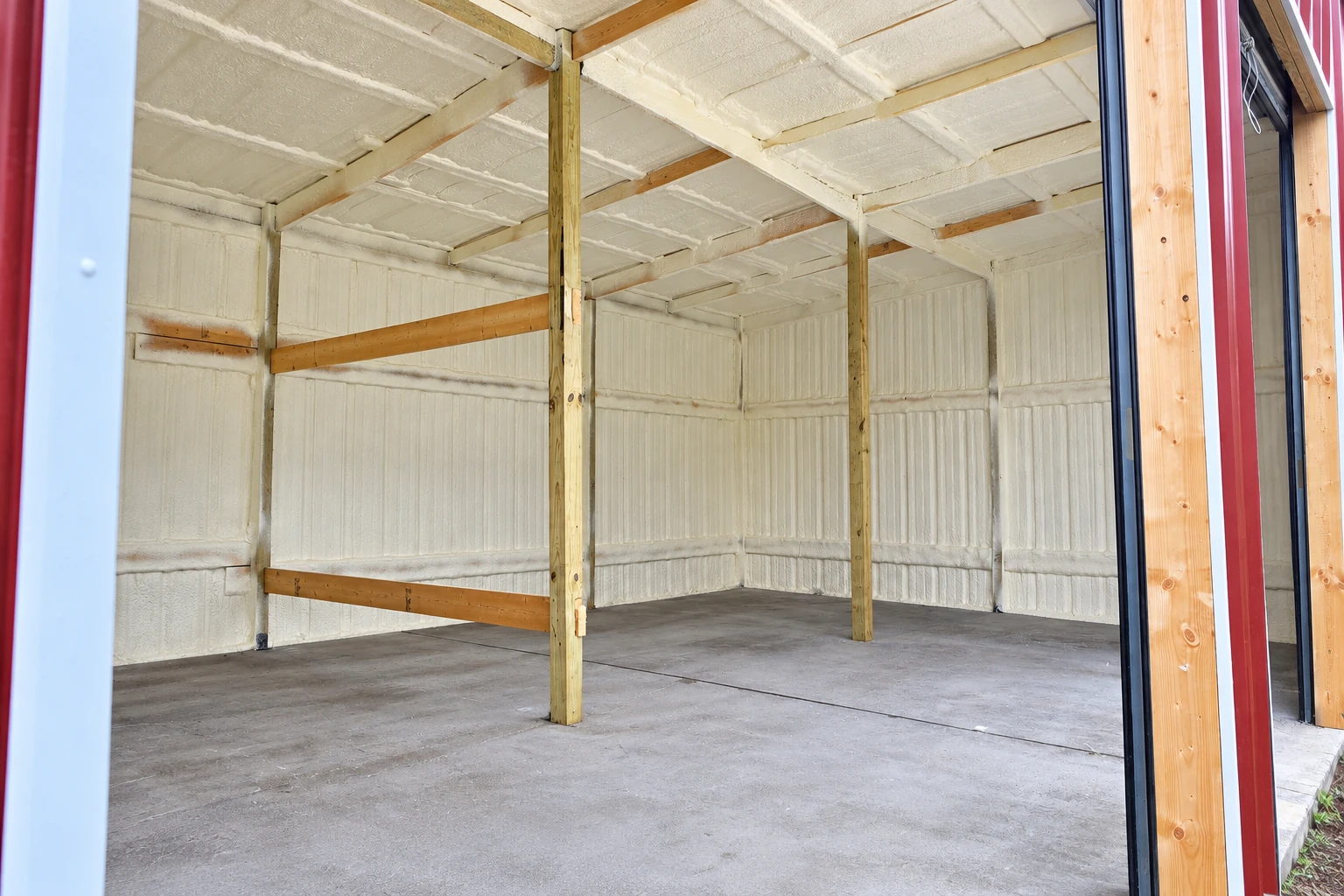 Garage interior with white spray foam insulation covering the walls and ceiling between exposed wooden framing.